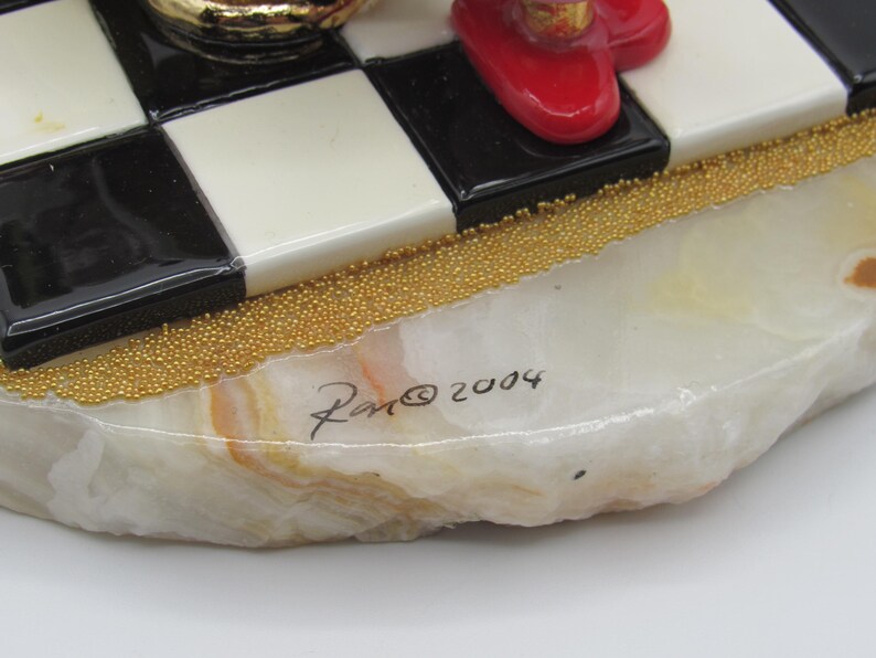 K&ouml;nnte beinhalten: Dekorative Figurenbasis mit einem schwarz-wei&szlig;en Schachbrettmuster. Die Basis besteht aus einem hellfarbigen Stein mit goldfarbenen Perlenakzenten. Die Signatur "Ron &copy; 2004" ist auf der Basis sichtbar.