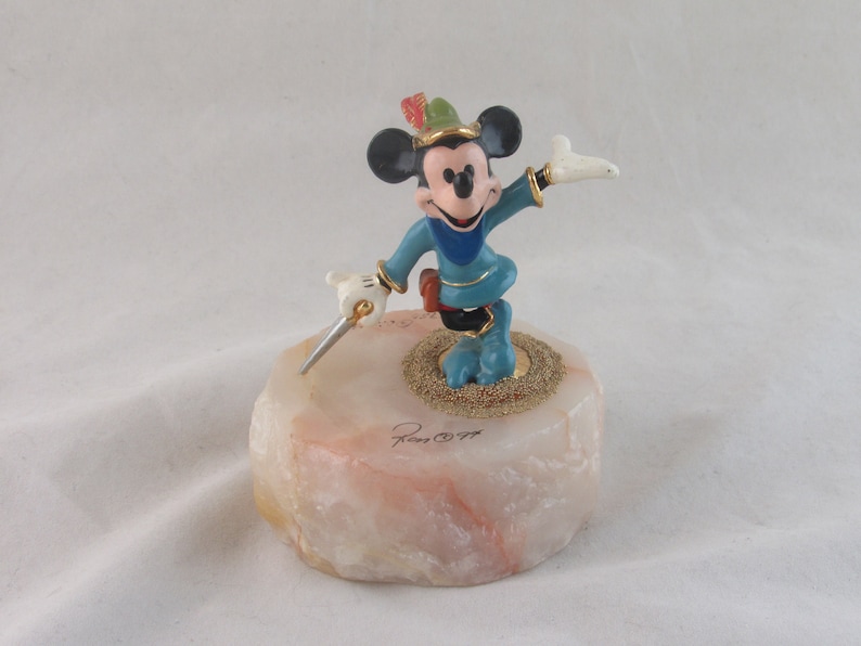 K&ouml;nnte beinhalten: Eine Mickey Mouse-Figur im Robin Hood-Kost&uuml;m, die ein Schwert h&auml;lt. Die Figur steht auf einem hellen, strukturierten Sockel mit goldenen Akzenten. Mickey tr&auml;gt einen gr&uuml;nen Hut mit Feder und ein blaues Outfit.