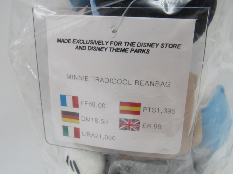 May include: A white tag with black text that reads "Made exclusively for the Disney Store and Disney Theme Parks" and "Minnie Tradicool Beanbag". The tag also shows prices in different currencies: FF69.00, DM18.50, LIRA21,000, PTS1,395, and &pound;6.99.