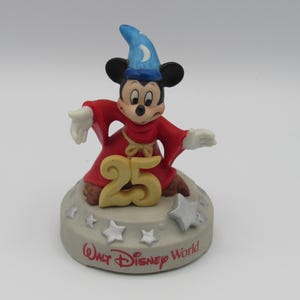 May include: A ceramic figurine of Mickey Mouse dressed as a sorcerer, wearing a red robe and a blue hat with a crescent moon. The figurine is on a base with the number "25" and the words "Walt Disney World".