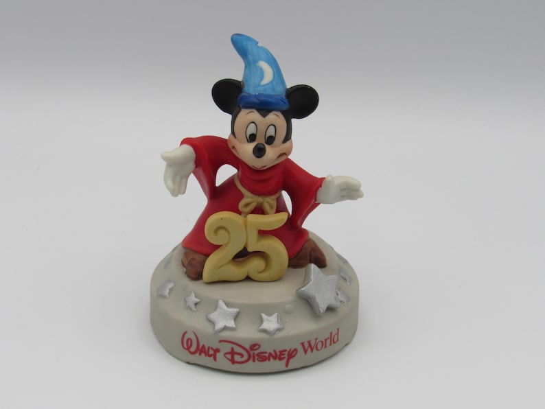 May include: A ceramic figurine of Mickey Mouse dressed as a sorcerer, wearing a red robe and a blue hat with a crescent moon. The figurine is on a base with the number "25" and the words "Walt Disney World".