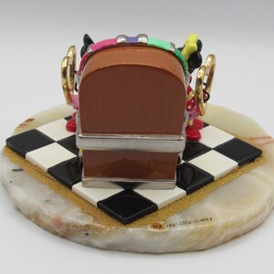 K&ouml;nnte beinhalten: Dekorative Figur mit einem braunen Jukebox-Design mit silbernen Akzenten, auf einer schwarz-wei&szlig; karierten Basis. Die Basis befindet sich auf einem hellfarbigen, oval geformten Stein. Die Figur hat farbenfrohe Details und goldfarbene Akzente.