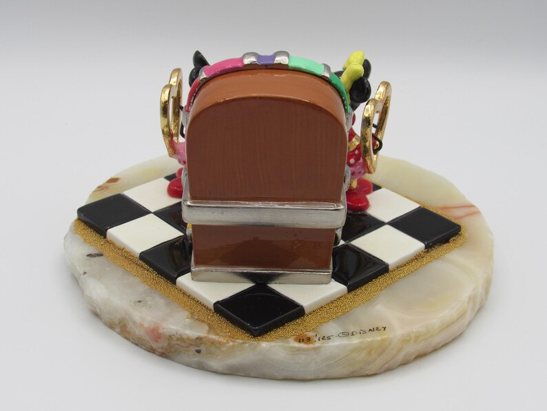 K&ouml;nnte beinhalten: Dekorative Figur mit einem braunen Jukebox-Design mit silbernen Akzenten, auf einer schwarz-wei&szlig; karierten Basis. Die Basis befindet sich auf einem hellfarbigen, oval geformten Stein. Die Figur hat farbenfrohe Details und goldfarbene Akzente.