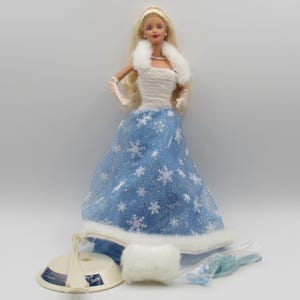 Puede incluir: Una muñeca rubia con cuello de piel blanca y falda larga azul con estampado de copos de nieve blancos y detalles brillantes. La muñeca lleva guantes blancos transparentes y un manguito blanco. Incluye un soporte beige redondo y un cepillo azul.