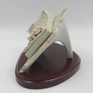 May include: A porcelain figurine of an open book with pink and purple flowers on the pages. The book is on a white stand with a brown wooden base.