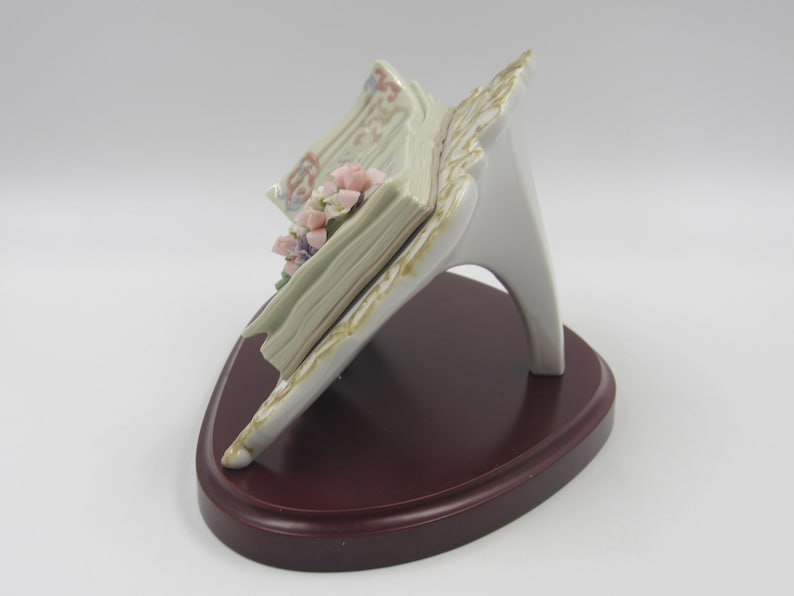 May include: A porcelain figurine of an open book with pink and purple flowers on the pages. The book is on a white stand with a brown wooden base.