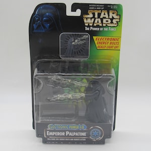 Könnte beinhalten: Eine Star Wars Actionfigur von Imperator Palpatine in versiegelter Verpackung. Die Verpackung zeigt das Star Wars-Logo, den Text "The Power of the Force" und den Satz "Electronic Energy Bolts Really Light Up!". Die Figur ist schwarz und hat dunkle Energieblitze.