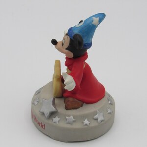 May include: A figurine of Mickey Mouse dressed as a sorcerer, wearing a blue hat with white stars and a red robe. The figure is standing on a gray base with white stars and the words "The World" in red.