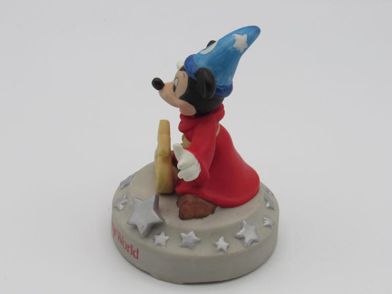 May include: A figurine of Mickey Mouse dressed as a sorcerer, wearing a blue hat with white stars and a red robe. The figure is standing on a gray base with white stars and the words "The World" in red.