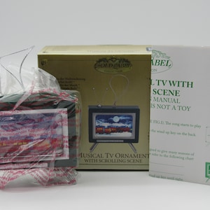 May include: A green musical TV ornament with a winter scene, packaged in plastic. The box reads "Gold Label Collection" and "Musical TV Ornament with Scrolling Scene". A manual is also included.