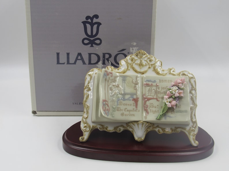 May include: A porcelain figurine of an open book with a bouquet of flowers on the right page. The book is on a wooden stand with ornate details. The figurine is made by Lladr&oacute;.