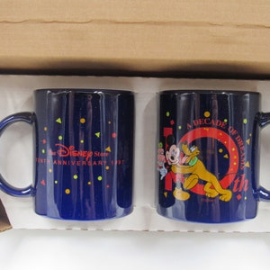 Puede incluir: Dos tazas de cerámica azules con un logotipo de Disney Store y un diseño de Mickey Mouse y Pluto. La taza de la izquierda dice "The Disney Store Tenth Anniversary 1987" y la taza de la derecha dice "A Decade of Dreams 10th".