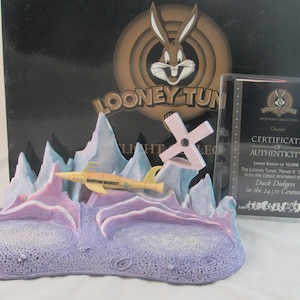 May include: A Looney Tunes collectible featuring a Martian Maggot spaceship, set against a backdrop of stylized mountains. The base is purple, with a yellow and blue spaceship. Includes a certificate of authenticity.