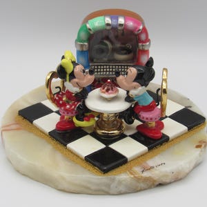 K&ouml;nnte beinhalten: Dekorative Figur mit Mickey und Minnie Mouse an einem Tisch mit einem Cupcake, vor einer Jukebox. Die Basis ist ein cremefarbener Stein mit einer schwarz-wei&szlig;en Schachbrettplattform. Die Jukebox hat einen bunten Bogen.