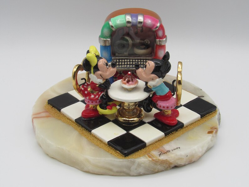 K&ouml;nnte beinhalten: Dekorative Figur mit Mickey und Minnie Mouse an einem Tisch mit einem Cupcake, vor einer Jukebox. Die Basis ist ein cremefarbener Stein mit einer schwarz-wei&szlig;en Schachbrettplattform. Die Jukebox hat einen bunten Bogen.