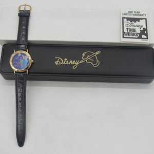 Könnte beinhalten: Eine goldfarbene Disney Time Works Armbanduhr mit blau-goldenem Zifferblatt und schwarzem Lederarmband. Die Uhr wird mit ihrer schwarzen Hülle präsentiert, die das Disney-Logo in Gold trägt. Eine weiße Box und eine Garantiekarte sind ebenfalls vorhanden.