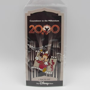 Könnte beinhalten: Disney-Anstecknadel mit Mickey Mouse in roter und weißer Kleidung, die eine Flagge mit dem Jahr 1999 hält. Die Anstecknadel ist in einer transparenten Plastikhülle mit dem Text "Countdown to the Millennium 2000" und "I Pin to Go" verpackt.