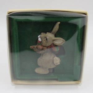 May include: A small, brown rabbit figurine with a red and green scarf, holding a brown stick in its paws. The figurine is displayed in a clear plastic box with a green velvet lining.