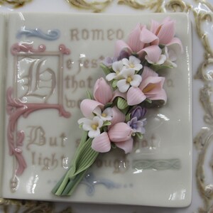 May include: A porcelain book with a pink and white floral bouquet on the cover. The book has a gold trim and the text "Romeo" is visible on the cover.