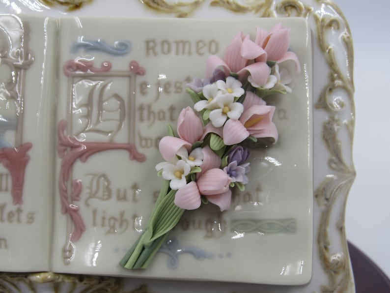 May include: A porcelain book with a pink and white floral bouquet on the cover. The book has a gold trim and the text "Romeo" is visible on the cover.