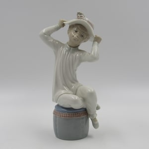 May include: A porcelain figurine of a child wearing a white dress and a white hat with pink flowers. The child is sitting on a gray and pink drum.