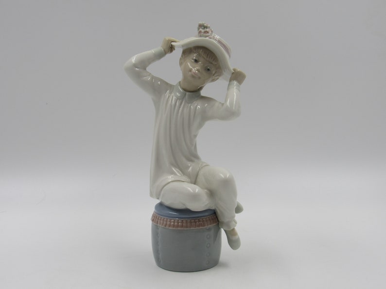 May include: A porcelain figurine of a child wearing a white dress and a white hat with pink flowers. The child is sitting on a gray and pink drum.