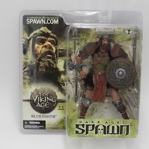 May include: A McFarlane Toys Dark Ages Spawn action figure in a clear plastic package. The figure is a Viking warrior with a shield, axe, and spear. The packaging includes the text "SPAWN.COM", "the VIKING AGE", and "BLUETOOTH".