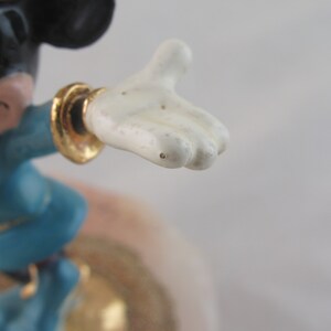 K&ouml;nnte beinhalten: Eine Mickey Mouse-Figur in einem blauen Outfit mit goldenen Akzenten. Die Figur ist mit einem ausgestreckten Arm dargestellt, der eine wei&szlig;e Hand zeigt. Der Hintergrund ist eine weiche, neutrale Farbe.