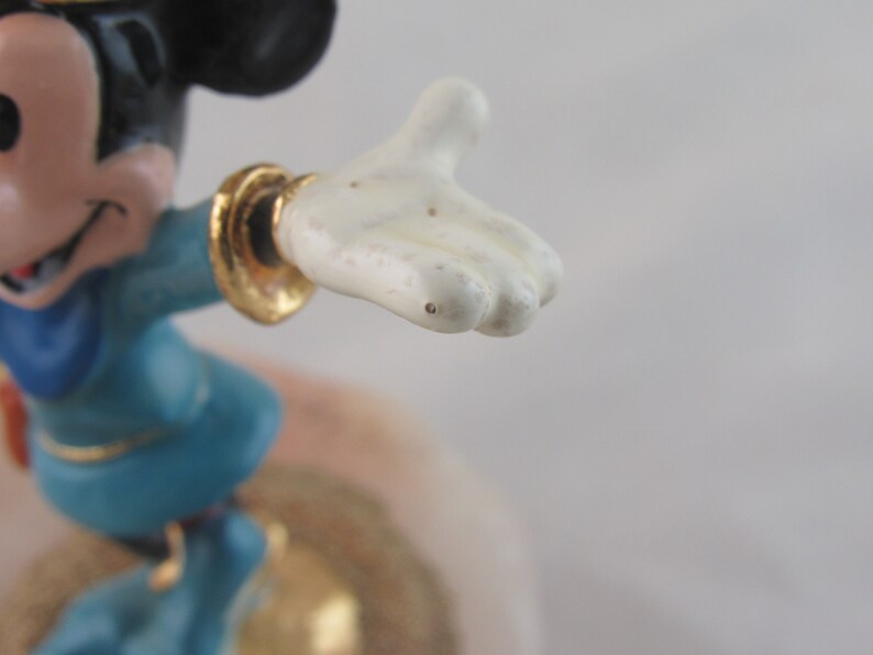 K&ouml;nnte beinhalten: Eine Mickey Mouse-Figur in einem blauen Outfit mit goldenen Akzenten. Die Figur ist mit einem ausgestreckten Arm dargestellt, der eine wei&szlig;e Hand zeigt. Der Hintergrund ist eine weiche, neutrale Farbe.