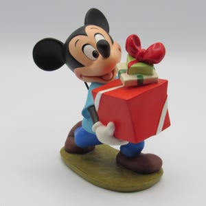 May include: A Mickey Mouse figurine holding a large red gift box with white ribbon and two smaller gifts. Mickey wears a blue shirt, blue pants, brown shoes, and white gloves. The figurine is on a green base.