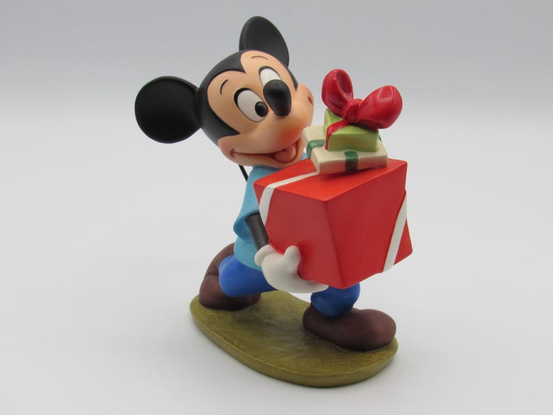 May include: A Mickey Mouse figurine holding a large red gift box with white ribbon and two smaller gifts. Mickey wears a blue shirt, blue pants, brown shoes, and white gloves. The figurine is on a green base.