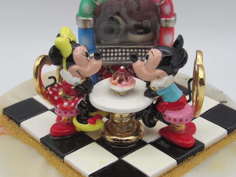 K&ouml;nnte beinhalten: Figur von Mickey und Minnie Mouse an einem Tisch mit einem Cupcake, vor einer Jukebox. Minnie tr&auml;gt ein rotes Polka-Dot-Kleid und Mickey ein blaues Hemd. Die Basis ist ein schwarz-wei&szlig;es Schachbrettmuster.