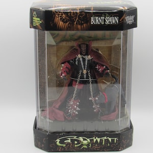 Puede incluir: Una figura de acción de Burnt Spawn dentro de una caja de exhibición hexagonal de plástico. El personaje viste un traje oscuro con capa roja, cadenas plateadas y armadura con pinchos, acompañado por una pequeña criatura gris.