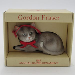 May include: A ceramic cat ornament in a box. The cat is gray and white with a red bow and a green collar. The box is tan with the words "Gordon Fraser" and "1985 Annual Dated Ornament" printed on it.