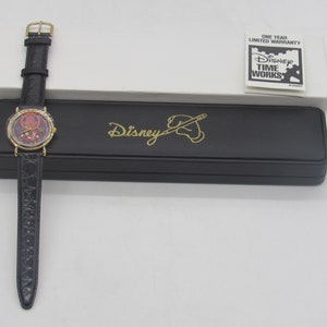 Könnte beinhalten: Eine goldfarbene Disney Time Works Uhr mit schwarzem Lederarmband und rundem Zifferblatt mit farbenfrohem Design. Die Uhr wird mit ihrer schwarzen Hülle präsentiert, die das Disney-Logo in Gold trägt. Eine Garantiekarte ist ebenfalls vorhanden.