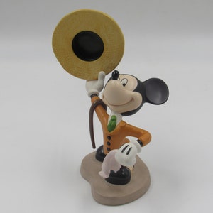 May include: A ceramic figurine of Mickey Mouse in a tan suit, green tie, and a straw hat. Mickey is holding the hat up with one hand and a cane in the other. The figurine is on a tan base.