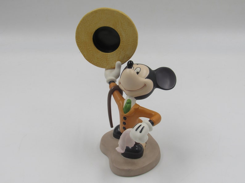 May include: A ceramic figurine of Mickey Mouse in a tan suit, green tie, and a straw hat. Mickey is holding the hat up with one hand and a cane in the other. The figurine is on a tan base.