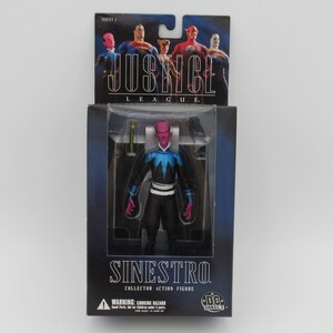 Op de afbeelding: Een Justice League-actiefiguur van Sinestro, een superschurk met een roze gezicht en een blauw-zwart kostuum. De figuur is verpakt in een doos met een afbeelding van de Justice League op de voorkant.
