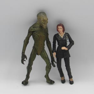 May include: Two action figures: a tall, green, reptilian alien and a woman in a black suit. The alien has a textured skin and a humanoid form. The woman has red hair and a badge. Both figures stand on a white surface.