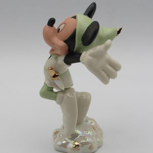Op de afbeelding: Een porseleinen beeldje van Mickey Mouse in een Peter Pan-ge&iuml;nspireerde outfit. Het beeldje is voornamelijk wit, met groene accenten op de hoed en mouwen. Gouden details sieren de outfit en de basis. Mickey staat met uitgestrekte armen.