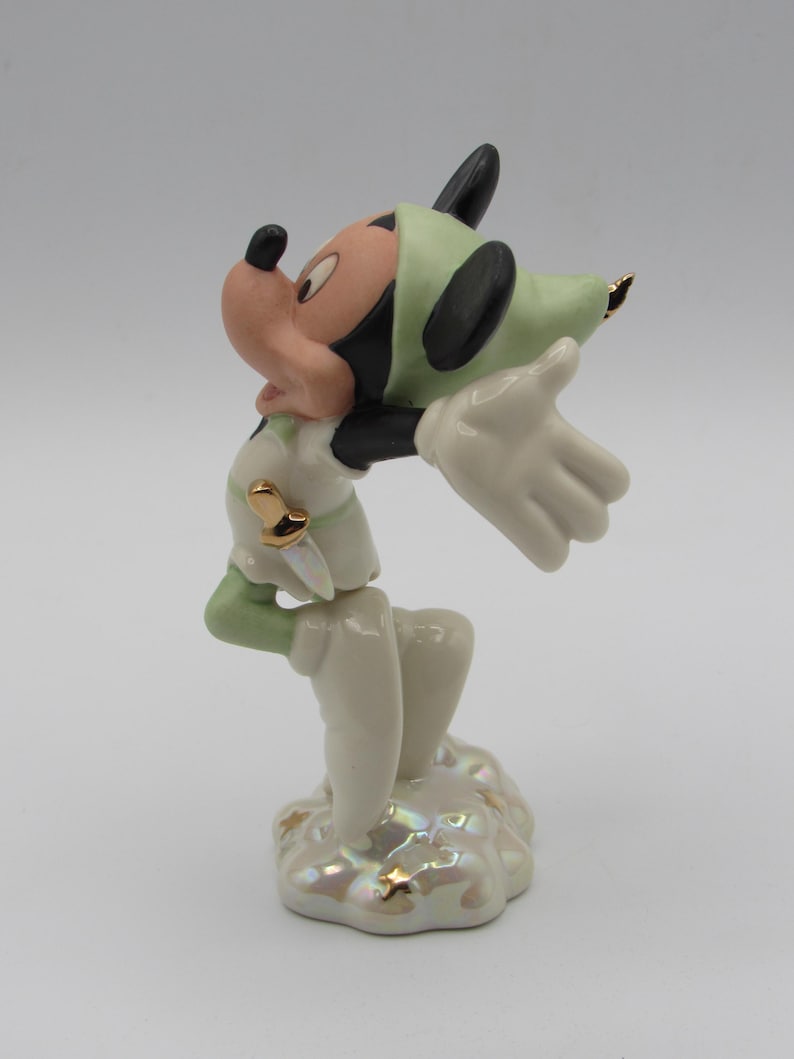 Op de afbeelding: Een porseleinen beeldje van Mickey Mouse in een Peter Pan-ge&iuml;nspireerde outfit. Het beeldje is voornamelijk wit, met groene accenten op de hoed en mouwen. Gouden details sieren de outfit en de basis. Mickey staat met uitgestrekte armen.