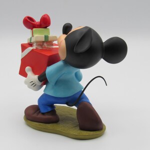 May include: A Mickey Mouse figurine carrying a stack of wrapped gifts. Mickey is wearing a blue shirt, blue pants, brown shoes, and white gloves. The gifts are red, green, and white with a red bow on top. The figurine is on a yellow base.