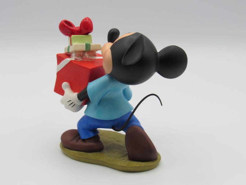 May include: A Mickey Mouse figurine carrying a stack of wrapped gifts. Mickey is wearing a blue shirt, blue pants, brown shoes, and white gloves. The gifts are red, green, and white with a red bow on top. The figurine is on a yellow base.