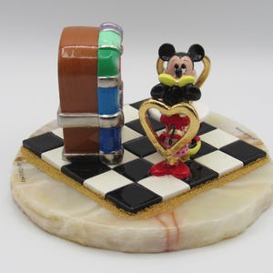 K&ouml;nnte beinhalten: Dekorative Figur mit einem braunen, blauen und gr&uuml;nen Jukebox-Design, mit einer Mickey Mouse Figur, die ein goldenes Herz h&auml;lt. Die Figur steht auf einer schwarz-wei&szlig; karierten Basis mit goldenem Rand, auf einer runden, cremefarbenen Steinbasis.