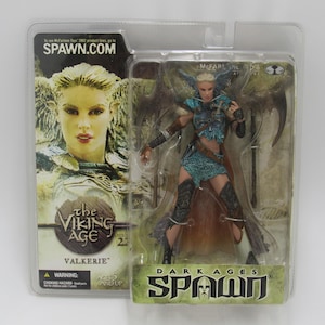 Op de afbeelding: Een Valkyrie actiefiguur uit de Dark Ages Spawn serie, verpakt in een doorzichtige plastic verpakking. Het figuur draagt blauwe en zwarte bepantsering, met een grote zeis. De verpakking bevat de tekst "The Viking Age" en "Spawn.com".
