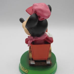 May include: A Minnie Mouse figurine in a red outfit and pink hat, riding in a red cart. The figurine is on a green base with the text "CLUB" in gold lettering. The figurine is a collectible item.