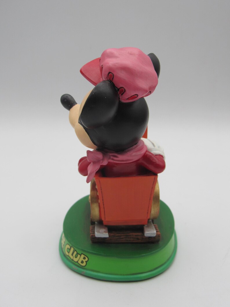 May include: A Minnie Mouse figurine in a red outfit and pink hat, riding in a red cart. The figurine is on a green base with the text "CLUB" in gold lettering. The figurine is a collectible item.