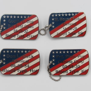 Può includere: Quattro targhette per cani in metallo con un disegno della bandiera americana. Ogni targhetta ha un campo blu con stelle bianche e strisce rosse e bianche. Il testo include nomi, gradi e gruppi sanguigni.