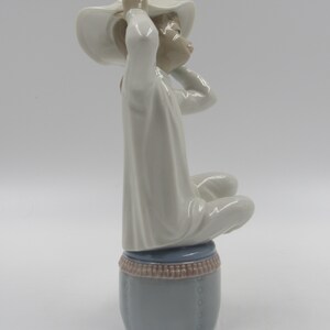 May include: A porcelain figurine of a girl wearing a white dress and a white hat with flowers. She is sitting on a blue and brown stool.