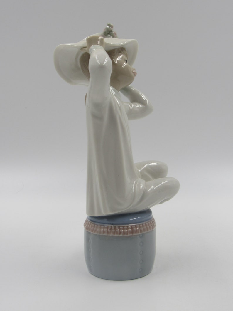 May include: A porcelain figurine of a girl wearing a white dress and a white hat with flowers. She is sitting on a blue and brown stool.